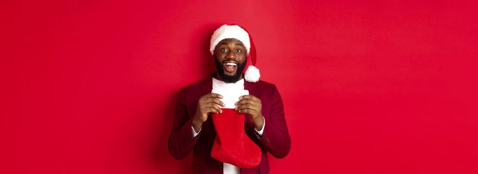 Excited black man open christmas sock with presents and sweets, smiling happy, standing in santa hat against red background.