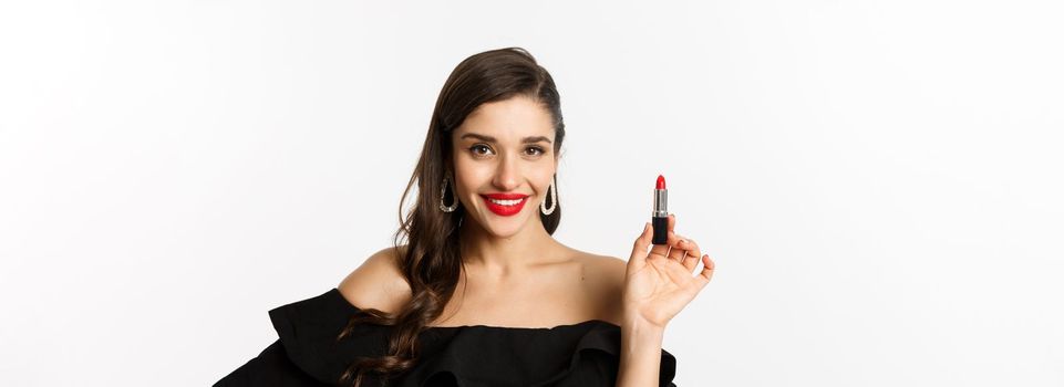 Fashion and beauty concept. Beautiful woman in black dress showing red lipstick and smiling, standing over white background. Copy space