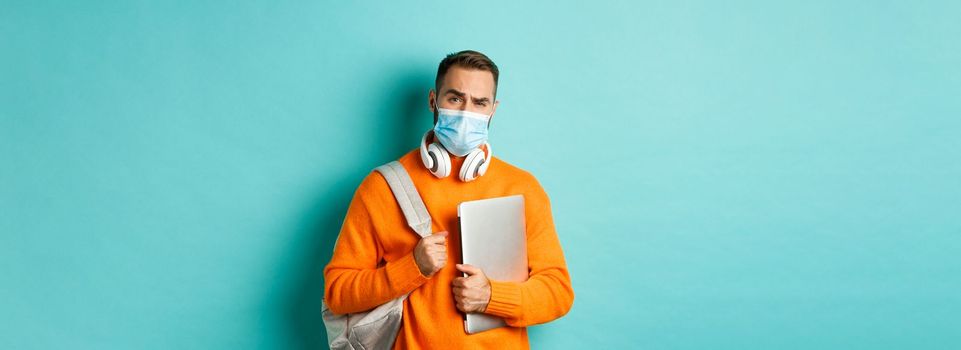 Handsome caucasian man with headphones and backpack, holding laptop and wearing medical mask, looking doubtful and skeptical, standing over light blue background.