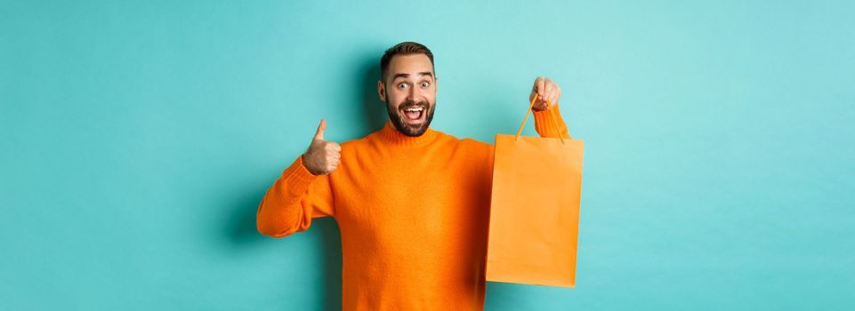 Happy man holding orange shopping bag and rejoicing from discount and celebrating, showing thumbs up and recommending, standing over blue background.