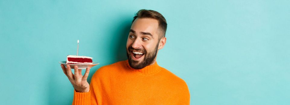 Close-up of happy adult man celebrating birthday, holding bday cake with candle and making wish, standing against turquoise background.