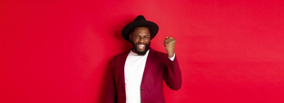 Fashion and party concept. Excited Black man rejoicing of winning, making fist pump gesture and say yes with satisfaction, standing against red background.
