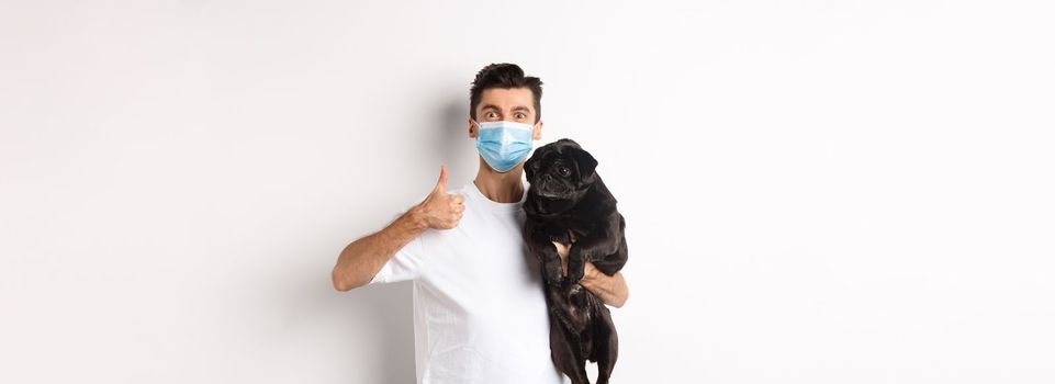 Covid-19, animals and quarantine concept. Young man in medical mask holding cute black pug dog, showing thumb up, like and approve, standing over white background.