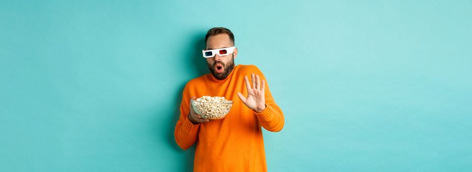 Image of man in 3d glasses watching movie, getting scared of special effects, looking in awe, standing with popcorn against white background.