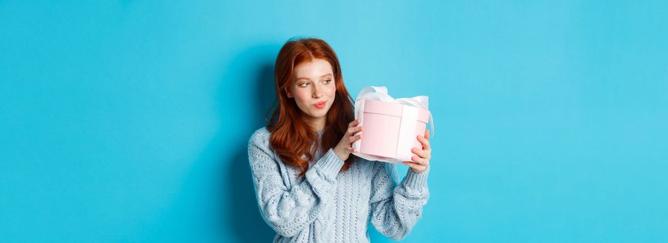 Curious teen girl with red hair, shaking gift box and wonder what inside, standing over blue background.