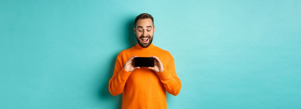 Excited guy demonstrating online store, showing mobile screen and looking amazed, standing over light blue background.