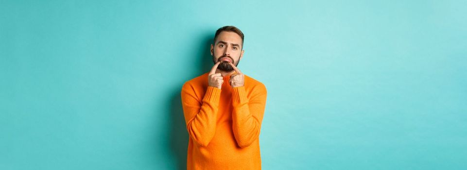 Image of gloomy bearded man, making sad face and frowning, standing upset in orange sweater against turquoise background.