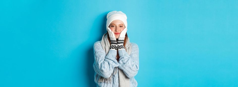 Winter and holidays concept. Cute girl in white beanie, sweater and gloves, squeezing cheeks and smiling pleased, warm-up after cold outdoors, standing over blue background.