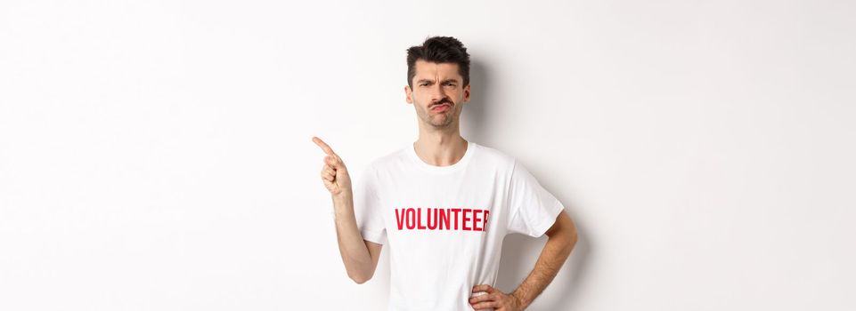 Skeptical and hesitant male volunteer in t-shirt grimacing doubtful, pointing finger left at promo offer, white background.