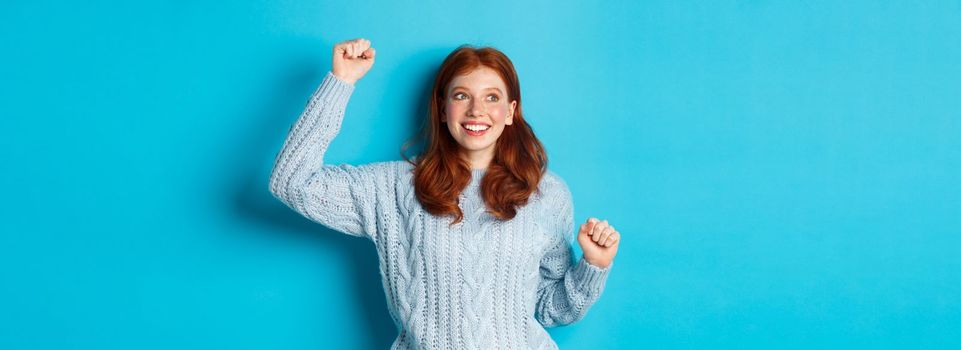 Happy redhead girl rooting for team, cheering with raised hand and smiling, celebrating victory or sucess, standing against blue background.