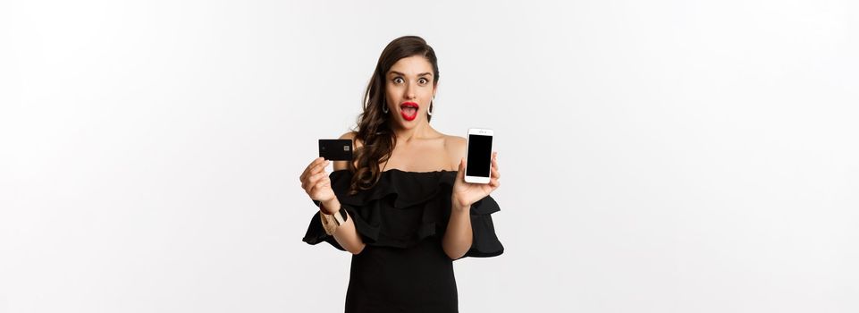 Fashion and shopping concept. Amazed beautiful woman showing smartphone screen and credit card, looking excited, buying online, white background.
