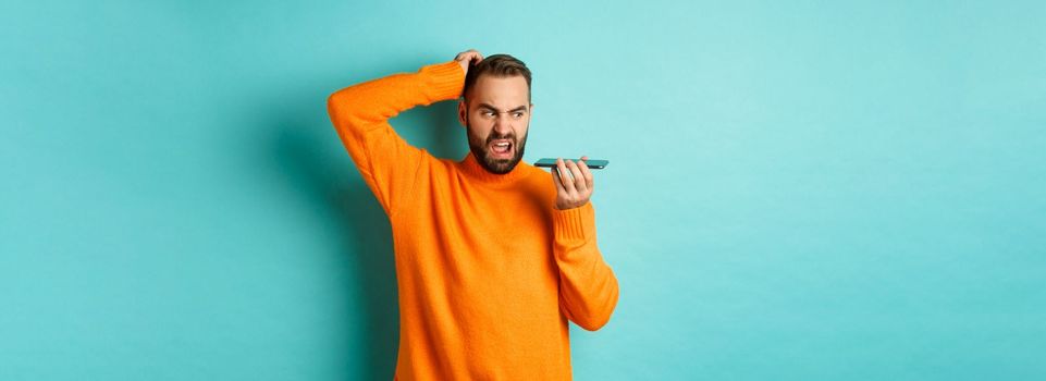 Confused man scratching head while talking on speakerphone, record voice message with indecisive face, standing in orange sweater over light blue background.