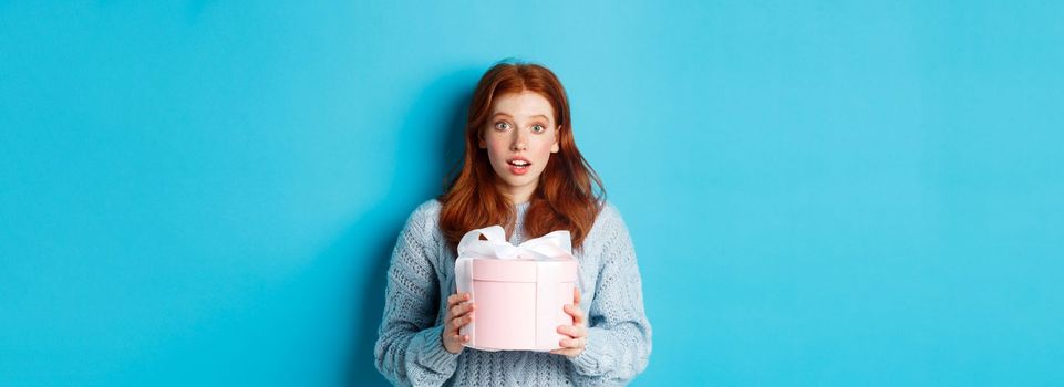 Surprised redhead girl receiving valentines gift, holding box with present and staring at camera amazed, wearing sweater, standing over blue background.