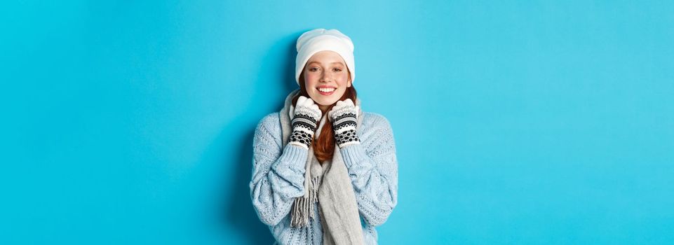 Winter and holidays concept. Cute redhead girl in white beanie and gloves smiling at camera, looking delighted, standing against blue background.