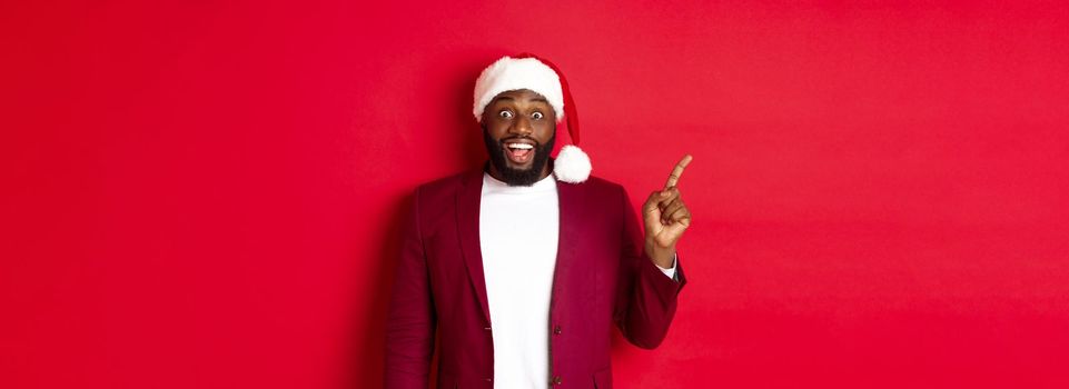 Christmas, party and holidays concept. Excited and amused Black man pointing finger left and smiling, showing logo, standing over red background.