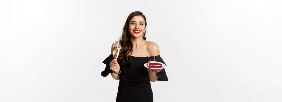 Celebration and party concept. Happy woman holding birthday cake and drinking champagne, smiling while standing in black dress against white background.