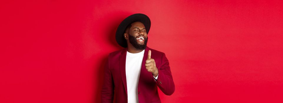 Handsome african american man having fun, showing thumb up and laughing from good joke, standing against red background.