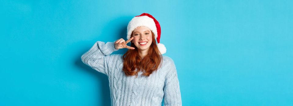 Winter holidays and Christmas Eve concept. Happy teenage girl with red hair, wearing santa hat, enjoying New Year, showing peace sign and smiling, standing over blue background.