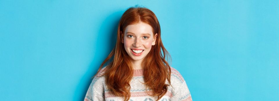 Close-up of cute redhead girl in sweater smiling happy at camera, standing against blue background.