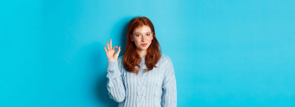 Confident redhead girl assuring you, showing okay sign and smiling, saying yes, approve and agree, standing over blue background.