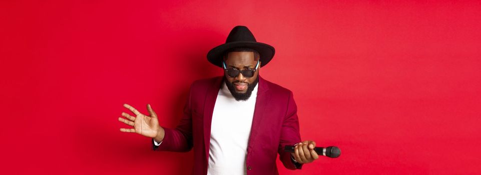 Passionate black male singer performing against red background, singing into microphone, wearing party outfit, standing over red background.