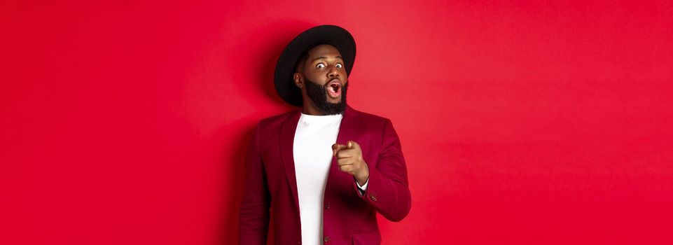 Shocked Black man gasping amazed and pointing finger at camera, recognize someone, standing in red blazer and hat against studio background.