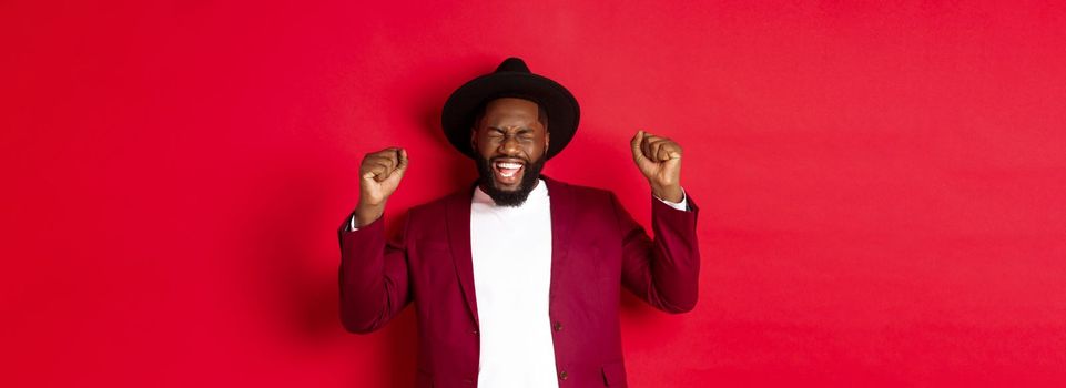 Fashion and party concept. Excited Black man winning prize, shaking raised hands and celebrating victory, partying New Year, standing against red background.