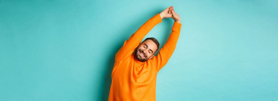 Image of handsome young man stretching hands and smiling after good rest, standing in orange sweater over light blue background.