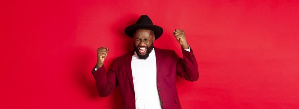 Successful Black man in suit saying yes, rejoicing of winning or achieving goal, raising hands up and screaming of joy, red background.