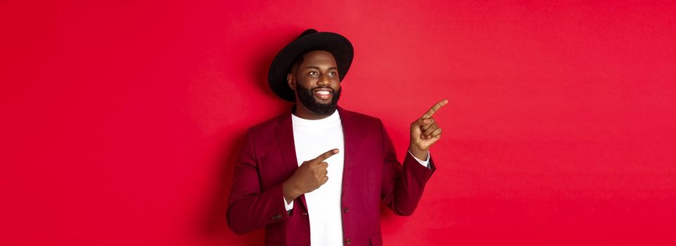 Winter holidays and shopping concept. Happy Black man pointing left and smiling, showing new year promo offer on red background.