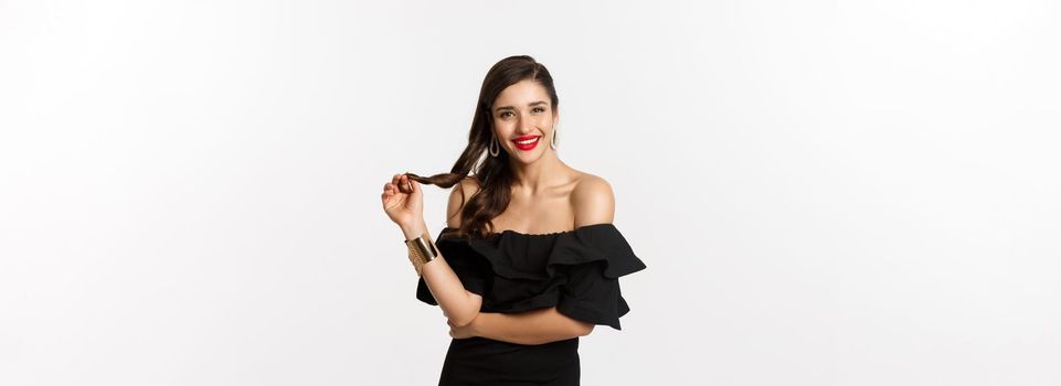 Beauty and fashion concept. Flirty woman in black dress, red lips, playing with hair and listening, looking at camera with interest, standing over white background.