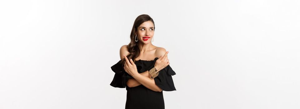 Fashion and beauty. Beautiful glamour woman in black dress, making choice, biting lip from temptation and pointing sideways, standing over white background.