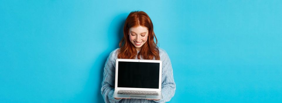 Cute redhead woman in sweater, showing and looking at laptop screen with pleased smile, demonstrating something online, standing over blue background.