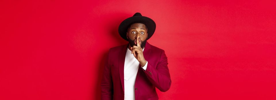 Fashion and party concept. African american man in classy hat hushing, asking keep secret, preparing surprise, standing over red background.