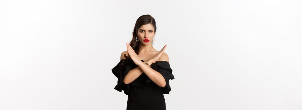 Fashion and beauty. Serious and confident female model in black dress, showing cross sign and frowning, stop gesture, telling no, standing over white background.