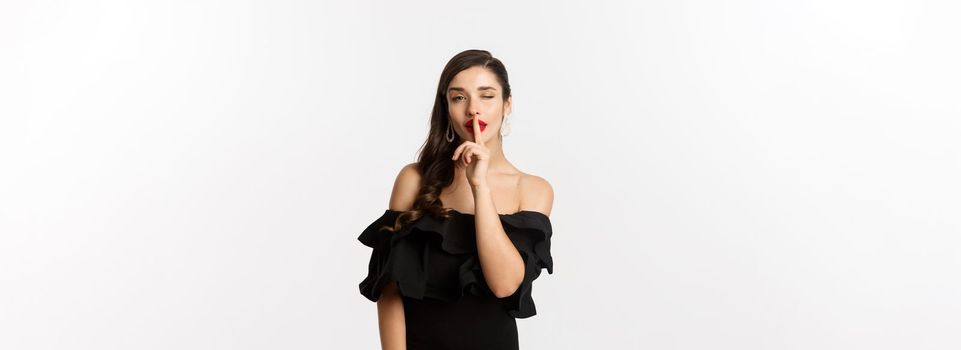 Fashion and beauty. Coquettish young woman in black dress, red lips, winking at camera and making hush sign, standing over white background.