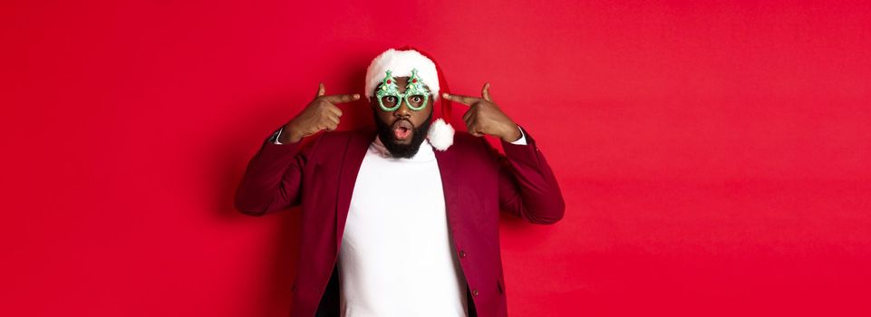 Merry Christmas. Cheerful Black man wearing funny party glasses and santa hat, smiling joyful, celebrating winter holidays, standing over red background.