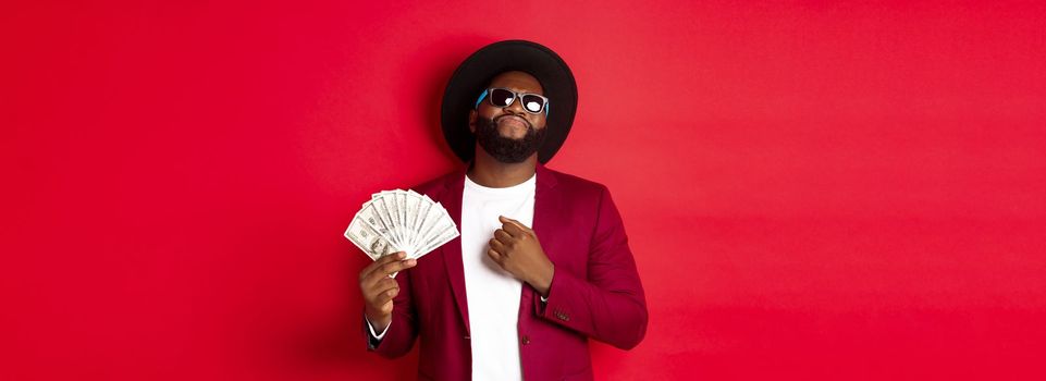 Sassy and cool african american man in sunglasses and hat, pointing at himself and showing dollars, looking confident against red background.