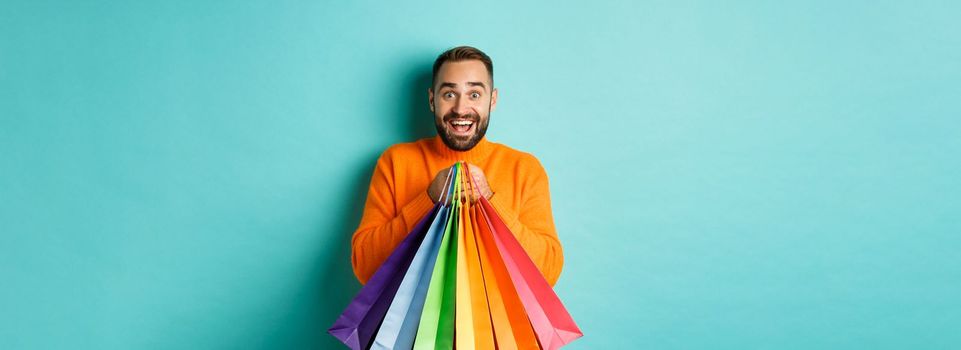 Excited adult man holding shopping bags and smiling, going to mall, standing over turquoise background.