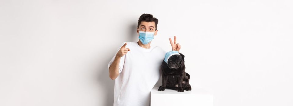 Covid-19, animals and quarantine concept. Happy dog owner and cute pug wearing medical masks, man pointing finger at camera and making funny bunny ears on pet, white background.