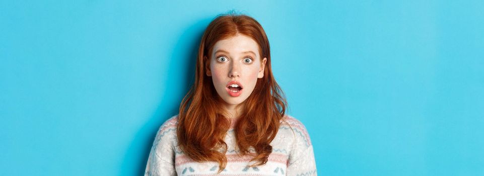 Close-up of shocked redhead girl drop jaw in awe, staring with amazement at camera, standing against blue background.