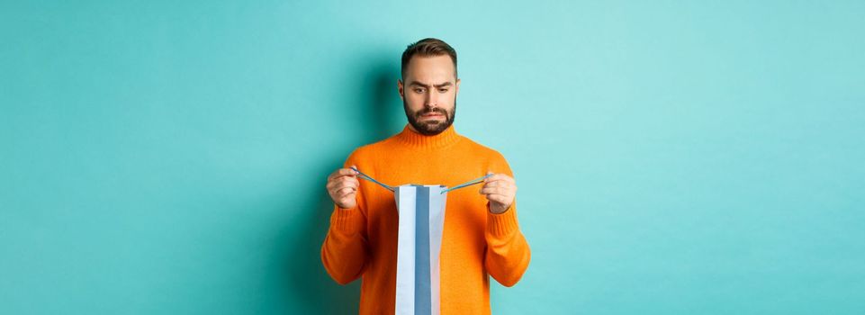 Disappointed man open shopping bag and dislike gift, frowning displeased, standing in orange sweater against turquoise background.