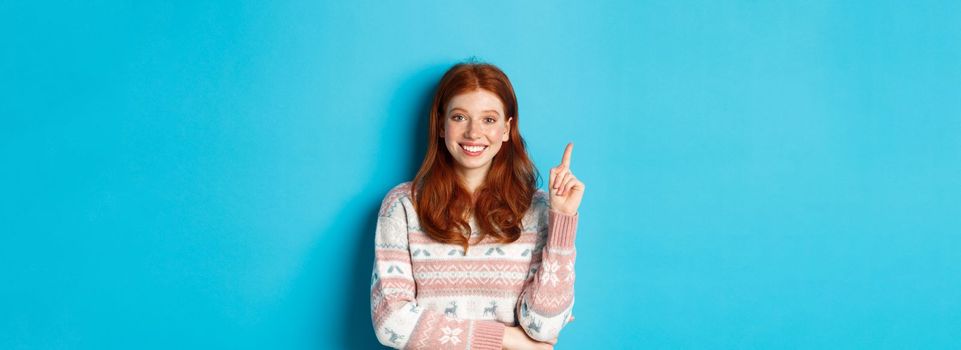 Excited redhead teen girl having an idea, raising finger and smiling, suggesting something, standing in sweater over blue background.