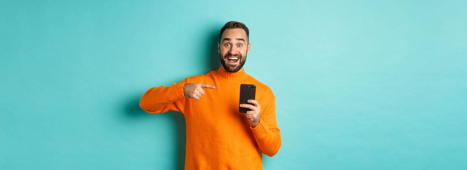 Excited man pointing at mobile phone, showing internet promo, standing over turquoise background, concept of online shopping.