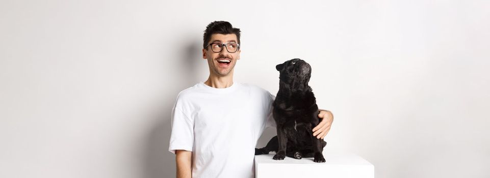 Amazed young man in glasses hugging his dog, pet owner and pug staring at upper left corner promo offer, standing over white background.
