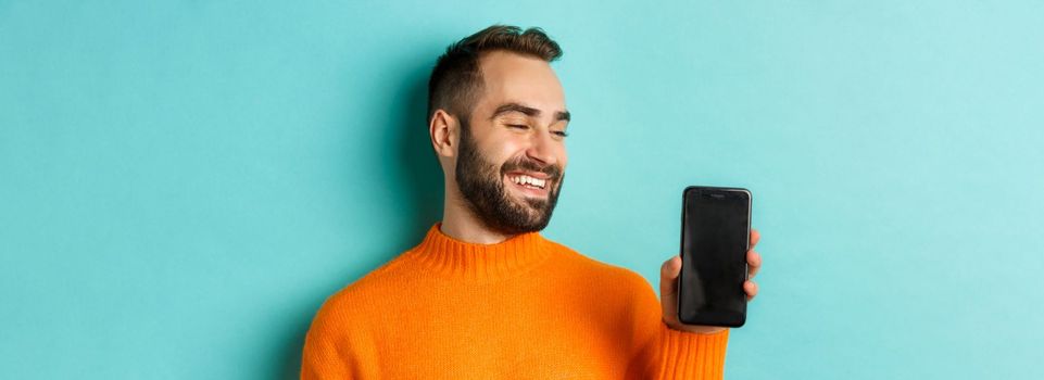 Close-up of young bearded man showing phone screen and looking satisfied, wearing orange sweater, standing against studio background.