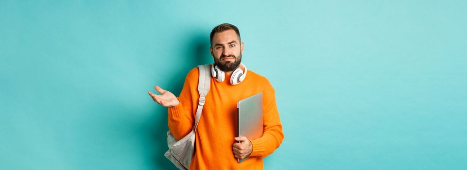 Image of troubled young man with headphones and backpack, shrugging confused and holding laptop, standing over light blue background.