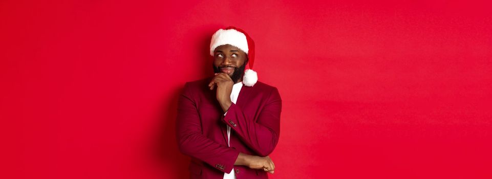 Christmas, party and holidays concept. Smiling african american man making new year plans, looking thoughtful at upper left corner, wearing santa hat, red background.