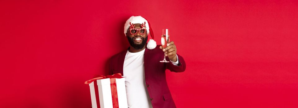 Christmas. Handsome african american man in party glasses and santa hat, holding new year gift and glass of champagne, wishing happy holidays, red background.