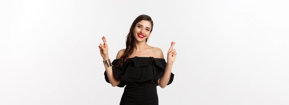 Fashion and beauty. Stylish glamour woman in black dress, red lips, looking optimistic and smiling while cross fingers, making wish, standing over white background.